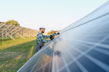 İki mühendis fotovoltaik güneş panellerini büyük bir güneş çiftliğine kuruyor ve kabloluyor, yeşil enerji üretiyorlar.