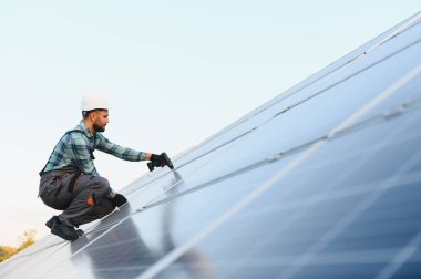 Erkek işçi, başlık ve eldiven takarak çatıdaki güneş paneline yerleştirme yapıyor ve temiz enerjiden faydalanıyor.