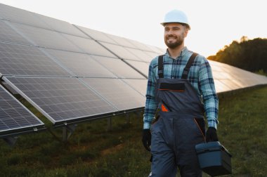Erkek mühendis, başlık takıyor ve tulum giyiyor güneş enerjisi santralinde yürüyor, bakım için bir alet çantası taşıyor.