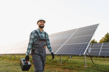 Sert şapkalı ve iş üniformalı teknisyen büyük bir güneş panelinde yürüyor, bakım için bir alet çantası taşıyor.