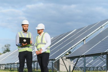 İki güvenlik ekipmanı mühendisi yenilenebilir enerji santralinde büyük güneş panellerinin yanında dururken bir dizüstü bilgisayarda verileri gözden geçiriyorlar.