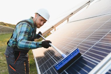 Sert şapkalı ve iş kıyafetli erkek teknisyen bakım yapıyor, cam sileceği ile güneş panelini siliyor.