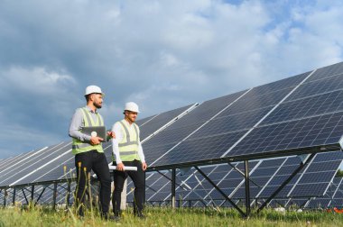 Yenilenebilir enerji mühendisleri güvenlik teçhizatlarıyla bulutlu bir gökyüzünün altındaki güneş panellerini inceleyerek sürdürülebilir teknolojiyi sembolize ediyorlar.