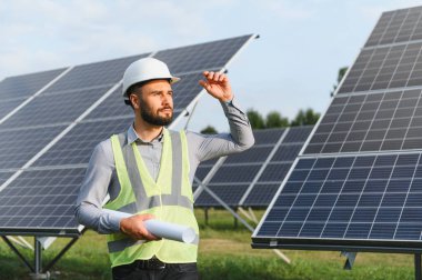 Sert şapka ve güvenlik yeleği giyen erkek mühendis güneş panellerini inceliyor, sürdürülebilir bir elektrik santralinin planlarını tutuyor.