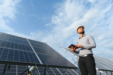 Erkek mühendis, başlık takıyor, elinde tablet tutuyor ve bir elektrik santralindeki güneş panellerini kontrol ediyor. Sürdürülebilir teknolojiye odaklanma