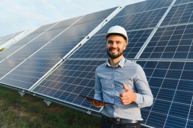 Başlık takan ve elinde tablet tutan mühendis güneş panellerinin önünde baş parmağını kaldırıyor.
