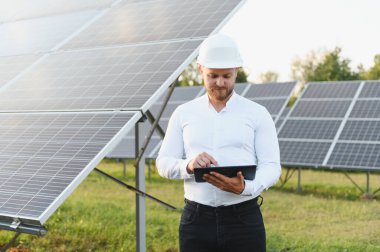 Sert şapkalı adam büyük güneş enerjisi santrali alanını denetliyor, yenilenebilir enerji sistemi performansını tabletten izliyor.