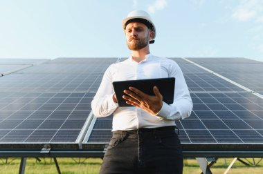 Güneş paneli dizisini denetleyen kasklı adam veri analizi ve yenilenebilir enerji yönetimi için tablet kullanıyor.