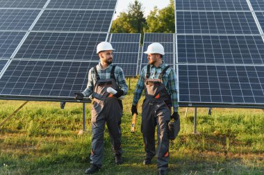 Güvenlik kaskı ve tulumu takan mühendisler, güneş paneli sıralı bir alanda yürürken planlarını tartışıyorlar.