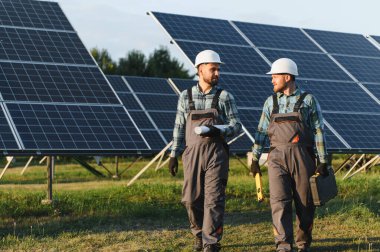 İki işçi güneş paneli alanında yürürken planlarını tartışıyor, yeşil enerji çözümleri geliştiriyor.