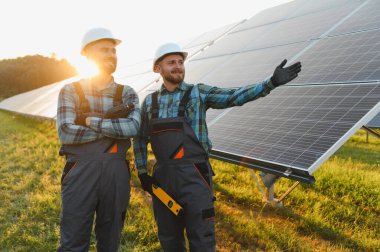 Günbatımında yenilenebilir enerji santraline güneş panelleri kuran ve denetleyen iki erkek mühendis, takım çalışması ve sürdürülebilir teknoloji