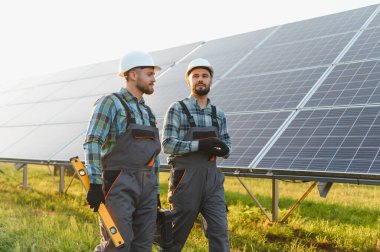 İki enerji işçisi, büyük bir dizi güneş panelinin yanında dikilip yeşil şapka ve tulum giyerek yeşil kariyerlerini destekliyor.