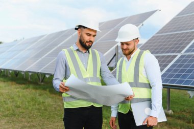 Güvenlik miğferleri ve yelekleri olan iki erkek mühendis yenilenebilir enerji sahasında büyük güneş panellerinin yanında dururken planlarını gözden geçiriyorlar.
