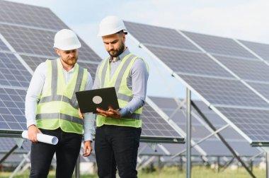 Güvenlik yelekleri ve miğferleri olan iki erkek mühendis güneş enerjisi projesinin verilerini güneş enerjisi tesis alanındaki bir dizüstü bilgisayardan inceliyor.