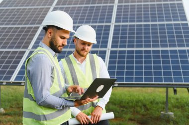 Güvenlik miğferi ve yeleklerinde çalışan iki mühendis güneş enerjisi verilerini bir güneş çiftliğindeki dizüstü bilgisayarda inceleyerek temiz yenilenebilir enerjiyi teşvik ediyorlar.