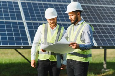Güvenlik yelekleri ve miğferler üzerinde çalışan iki mühendis bir güneş enerjisi çiftliğinin planlarını gözden geçirip takım çalışmasını ve sürdürülebilir gücü sembolize ediyorlar.