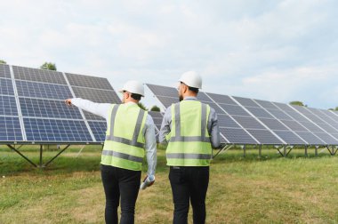 Güvenlik yelekleri ve miğferlerinde çalışan iki mühendis, yenilenebilir enerji ve sürdürülebilirliği vurgulayan güneş paneli kurulumunu inceler ve tartışır