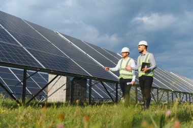 Yeşil alanda güneş panellerini denetleyen güvenlik yelekleri ve miğferler giyen iki erkek mühendis, temiz yenilenebilir enerji büyümesini sembolize ediyor.