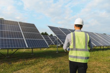 Büyük bir fotovoltaik güneş çiftliği sahasındaki güneş panellerini incelerken elinde planlar tutan çelik yelek mühendisi.