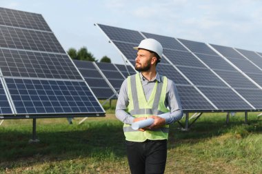 Sıkı şapka ve güvenlik yeleği giyen profesyonel mühendis, güneş paneli alanını incelerken elinde planlar tutuyor.