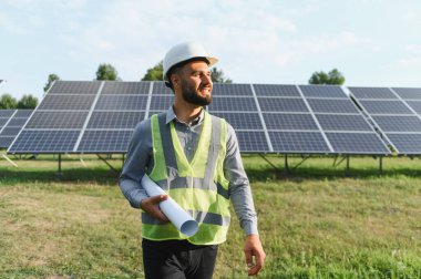 Başlık ve güvenlik yeleği giymiş mühendis elinde planlar, güneş panellerinin önünde dururken gülümsüyor.