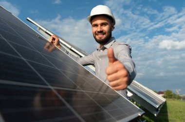 Fotovoltaik dizinin yanında durup, temiz ve sürdürülebilir enerji çözümlerine onay veren, sert şapkalı gülen bir işçi.