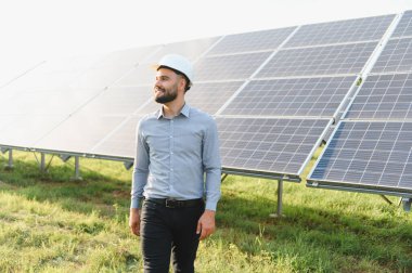 Erkek mühendis, güneş panellerinin yanında yürürken, yenilenebilir enerji ve çevresel sürdürülebilirliği sembolize ediyor.