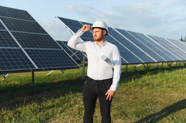 Mühendis sabit bir şapka takıyor ve elinde bir planla yenilenebilir enerji tesisinde güneş panellerinin önünde duruyor.