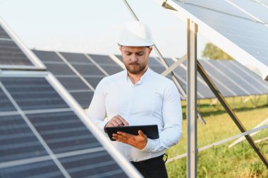 Sert şapka takan mühendis, yenilenebilir enerji ve yeşil teknolojiyi sembolize eden bir tablette güneş paneli performansını kontrol ediyor.