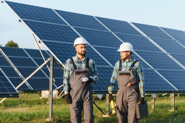 Sert şapka ve tulum giymiş mühendisler yenilenebilir enerji güneş enerjisi çiftliğindeki güneş panellerini incelerken planlarını tartışıyorlar.