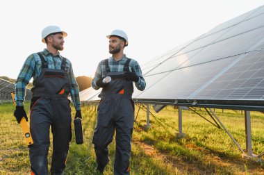 Güneş paneli alanında yürüyen iki erkek mühendis planlarını tartışıyor ve yenilenebilir enerji tesisatını yönetiyor.