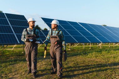 Güneş paneli kurulumu sırasında tarlada yürürken kasklı ve tulumlu iki işçi.