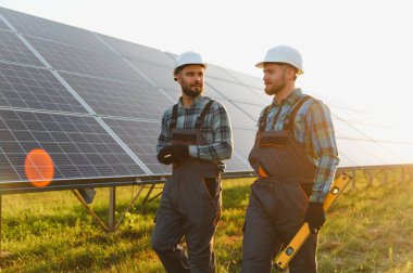 Güneş panellerini inceleyen, sürdürülebilir yenilenebilir enerji üretimi sağlayan, kalın şapkalı ve tulumlu iki işçi