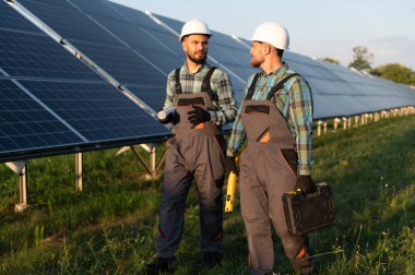 İki güneş enerjisi mühendisi iş tartışıyor, büyük bir güneş çiftliğinde sistem işleyişini kontrol ediyor, yeşil enerji çözümü sağlıyor