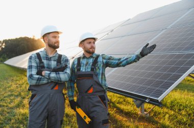Günbatımında güneş paneli alanında duran iki mühendis, biri sürdürülebilir enerji kurulumunu işaret ediyor.