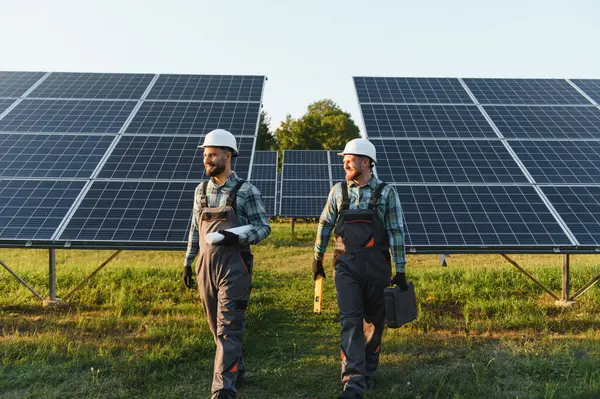Sert şapka ve tulum giyen iki mühendis güneş panellerinden geçerek yenilenebilir enerji üretiyorlar.