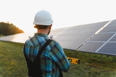 Mühendis sabit bir şapka takıyor ve bir seviyeyi tutuyor. Elektrik santralindeki güneş panellerini kontrol ediyor ve yeşil enerji üretiyor.