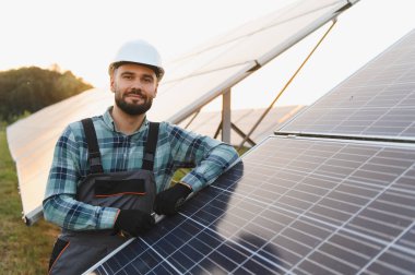 Erkek teknisyen, kasketli ve tulumlu. Sahadaki bir güneş panelini incelerken güvenle gülümsüyor.