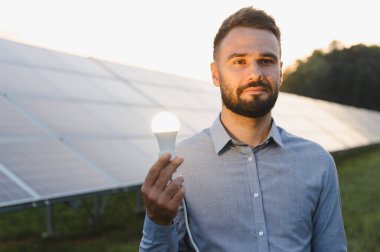 Güneş paneli önünde parlayan bir ampul tutan adam yenilenebilir enerji, sürdürülebilirlik ve yenilikleri temsil ediyor.