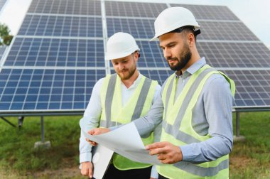 Güvenlik teçhizatı inceleme projesindeki iki mühendis yenilenebilir enerji tesislerinde büyük güneş panellerinin yanında dururken