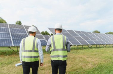 Güneş panellerinin sıralarını araştıran güvenlik yeleği ve miğfer mühendisleri, sürdürülebilir enerji gelişimini sembolize ediyorlar.
