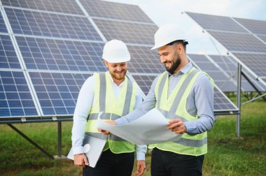 Güvenlik yelekleri ve miğferler üzerinde çalışan iki mühendis yenilenebilir enerji çözümleri planlayan bir güneş paneli tesis alanının planlarını gözden geçiriyorlar.
