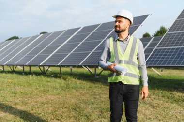 Sert şapka ve yansıtıcı yelek giyen mühendis güneş panellerinin önünde yuvarlanmış planlar tutuyor. Yenilenebilir enerji kavramı