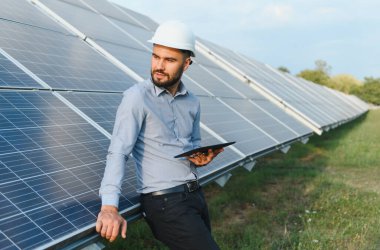 Bir tablet tutan, güneş panellerine yaslanan ve bir enerji santrali alanındaki sürdürülebilir enerji üretimini izleyen bir mühendis.