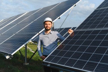 Beyaz şapka ve gömlek giyen erkek mühendis, güneş panelleri arasında gururla duruyor, yenilenebilir enerji ve sürdürülebilirliği sembolize ediyor.
