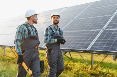 Sert şapka ve tulum giyen iki teknisyen, sürdürülebilir bir enerji sistemi kurmak için büyük bir güneş paneli dizisi üzerinde çalışıyorlar.