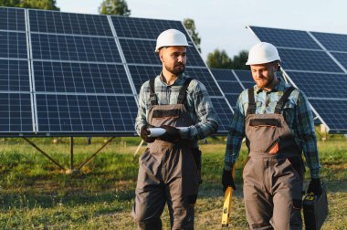 Güneş çiftliğinde yürüyen, baret ve tulum giyen işçiler ellerinde planlar ve aletler tutuyorlardı. Yenilenebilir enerji kavramı