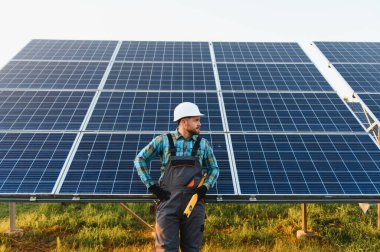 Sert şapka ve tulum giyen işçiler yenilenebilir enerji teknolojisini temsil eden büyük güneş panellerinin önünde güvenle duruyorlar.