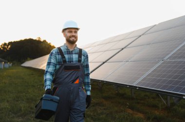 Kask ve tulum giymiş gülümseyen işçiler, alet çantası taşıyor, güneş enerjisi santralinde yürüyor, sürdürülebilir enerji sağlıyor.