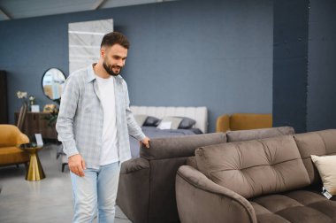 Genç adam gülümsüyor, evi için yeni bir kanepe seçiyor modern bir mobilya galerisinde seçenekleri araştırıyor.